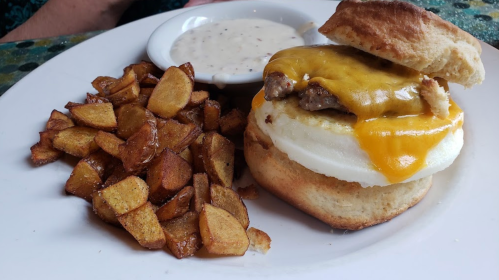 A plate with a biscuit sandwich filled with sausage and cheese, served with crispy potatoes and a side of gravy.